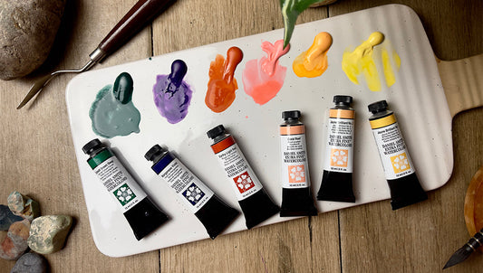 A photo of a white ceramic watercolour palette with 12 bright Opus Essential Watercolour paints are on a table with a colour chart, tubes of the colours, and a hand holding a paint brush that is being dipped into red paint in the palette.