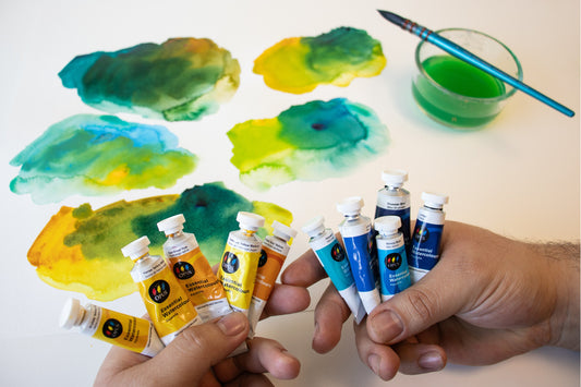 a number of paint swatches made of different blues and yellows in the background behind two hands - in one hand is 5 tubes of yellow watercolour and in the other hand is 5 tubes of blue watercolour