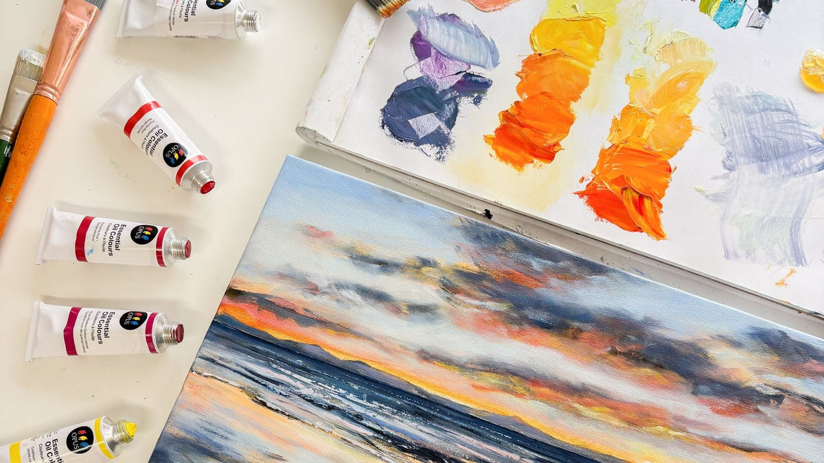 Flat lay of oil painting workspace with Opus Essential oil colour tubes, brushes, and palette showing warm yellows and oranges, alongside a finished sunset seascape painting with waves and glowing sky.