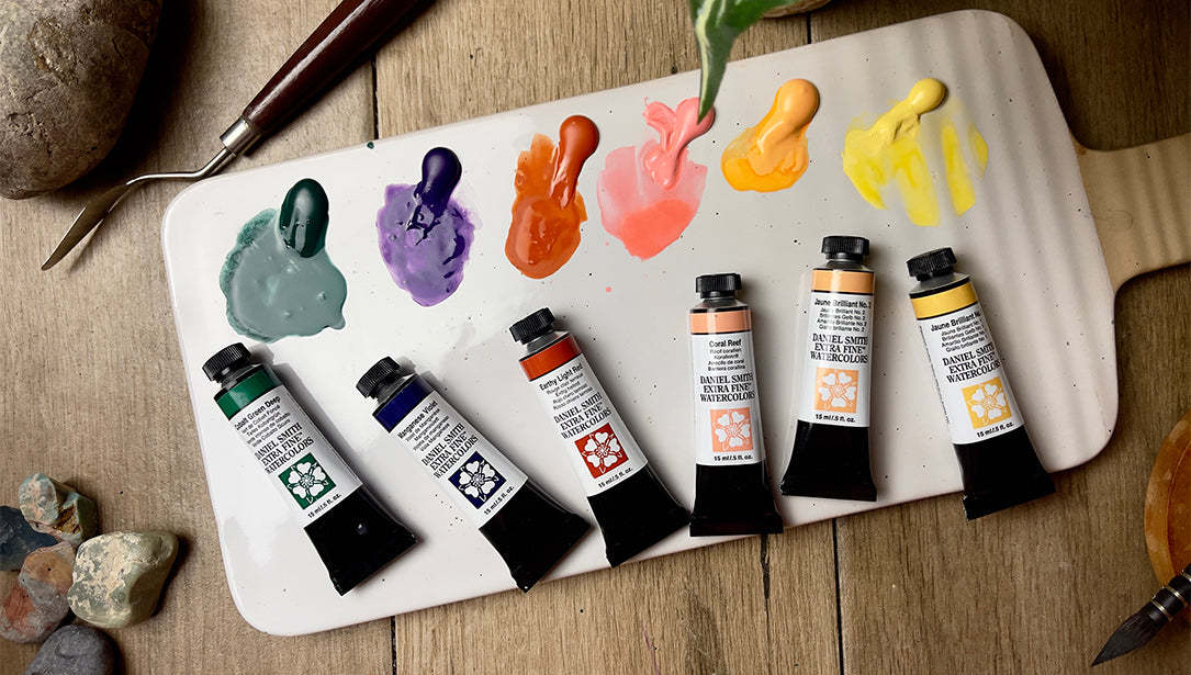 A photo of a white ceramic watercolour palette with 12 bright Opus Essential Watercolour paints are on a table with a colour chart, tubes of the colours, and a hand holding a paint brush that is being dipped into red paint in the palette.