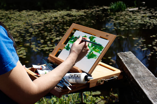 Artist painting outdoors beside a lily-covered pond using a portable plein air easel.