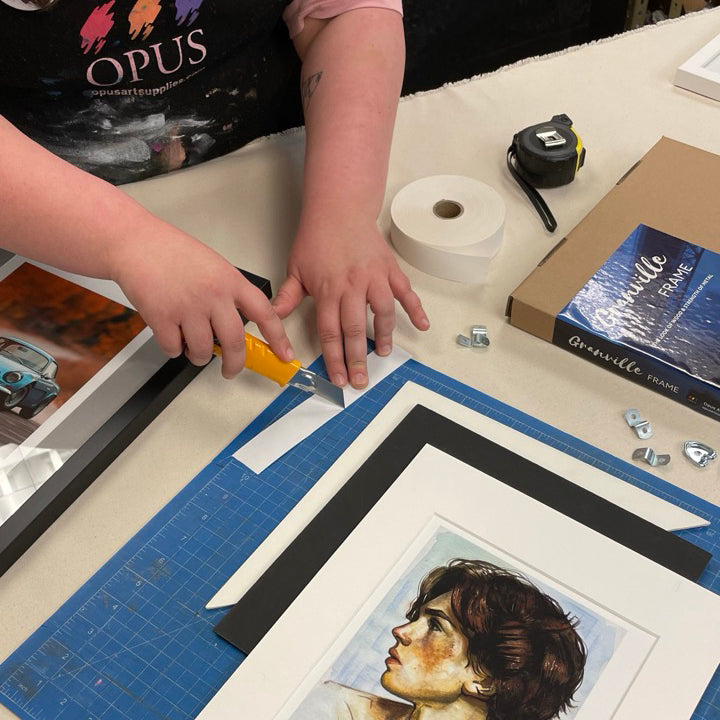 A woman's hands at a framing table, cutting a piece of linen tape with an Xacto Knife