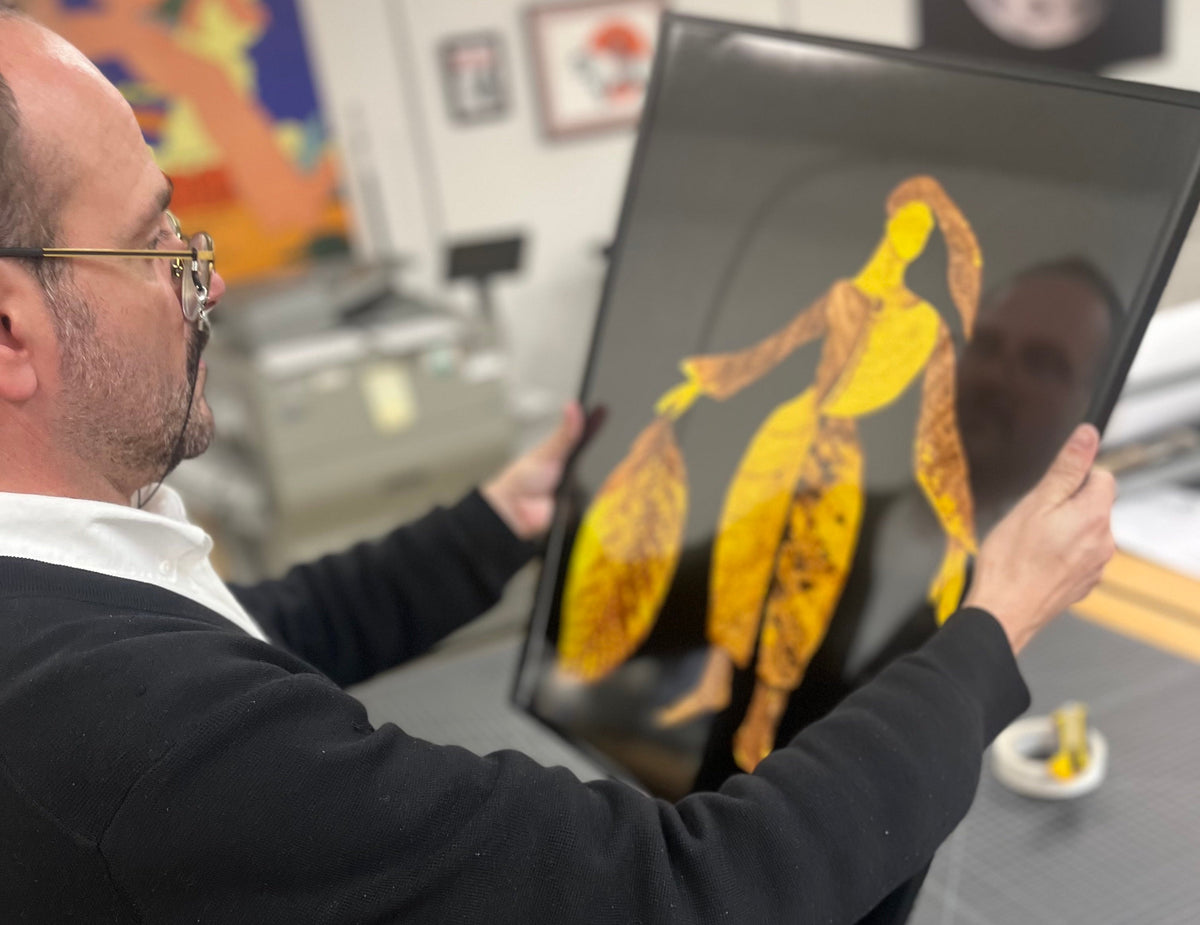 Opus Print Space team member holding a framed botanical fine art print by Sarah Chase, featuring a yellow figure composed of leaves against a black background.
