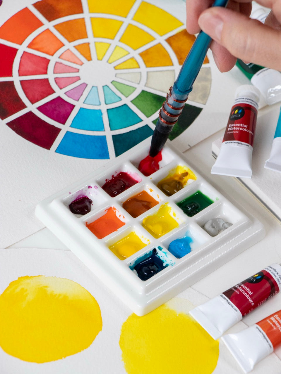 A photo of a white ceramic watercolour palette with 12 bright Opus Essential Watercolour paints are on a table with a colour chart, tubes of the colours, and a hand holding a paint brush that is being dipped into red paint in the palette.