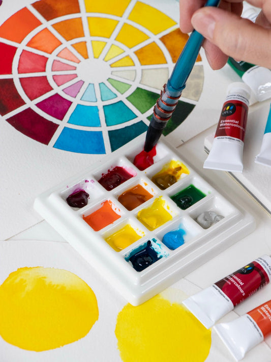 A photo of a white ceramic watercolour palette with 12 bright Opus Essential Watercolour paints are on a table with a colour chart, tubes of the colours, and a hand holding a paint brush that is being dipped into red paint in the palette.