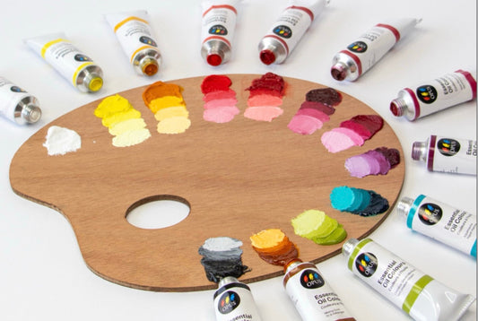 A wooden palette with all 12 of the new Opus Essential Oil Colours painted out with increasing amounts of white around the edge of the palette, with the corresponding colour's tube of paint on the white table, surrounding the palette.