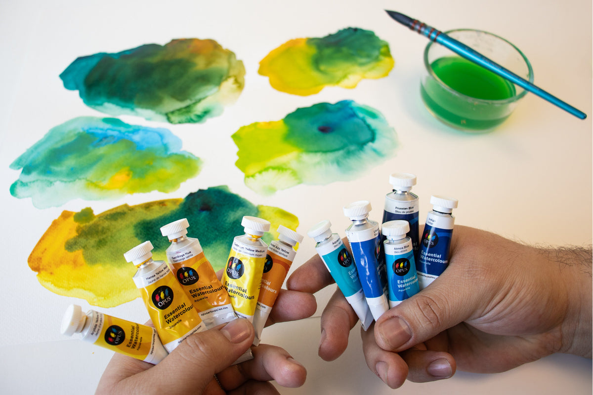 a number of paint swatches made of different blues and yellows in the background behind two hands - in one hand is 5 tubes of yellow watercolour and in the other hand is 5 tubes of blue watercolour