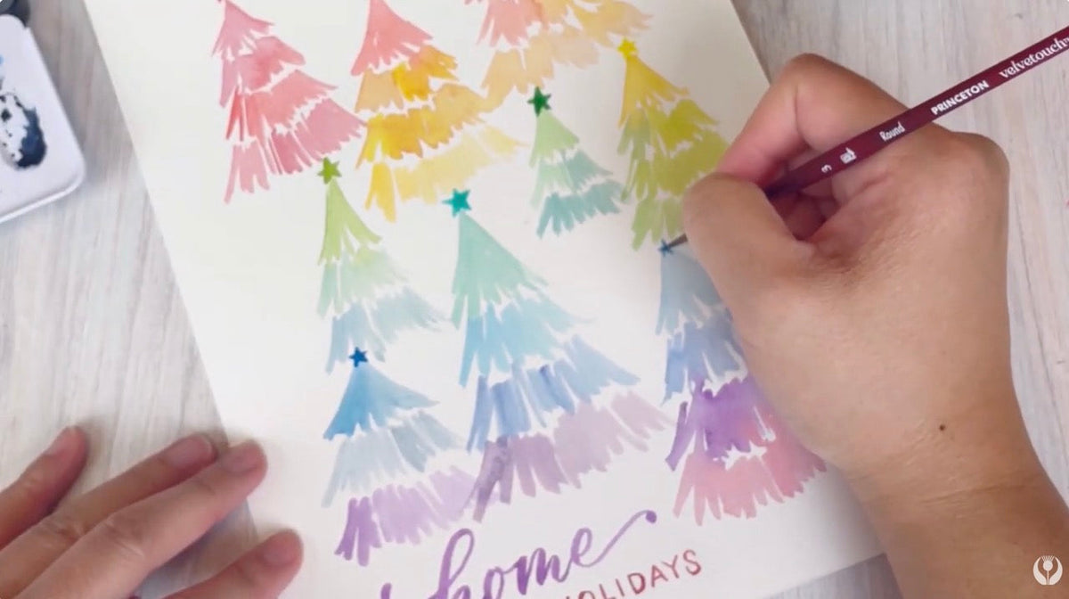 A watercolour painting of rainbow coloured holiday trees and the hand and brush of the artist