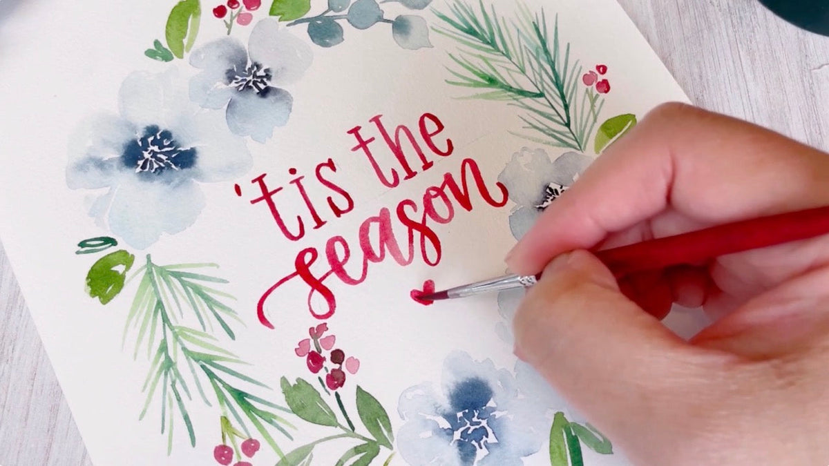 an artist's hand is painting a watercolour card with a wintery wreath with flowers and berries and the words tis the season