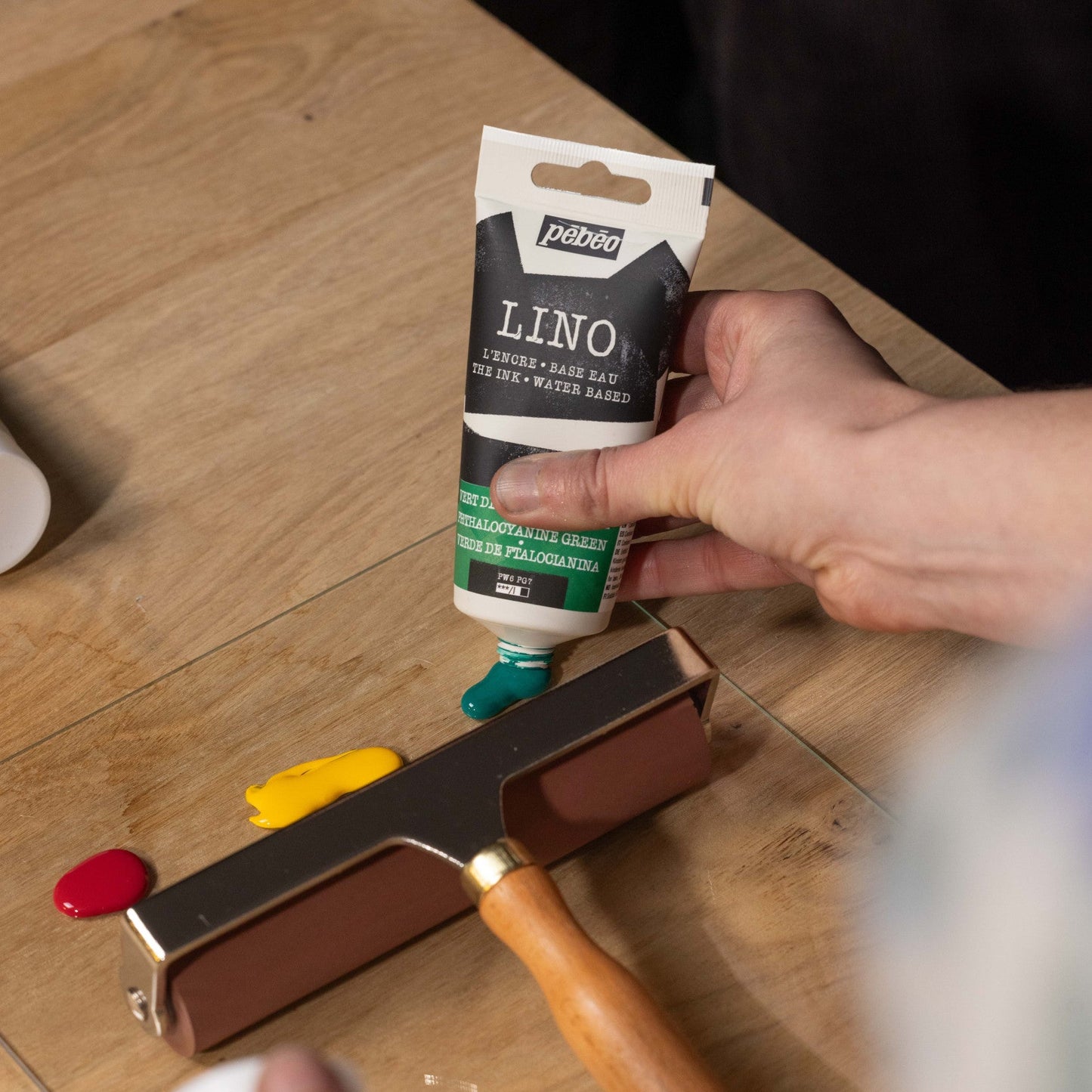 Person holding a tube of Pébéo Lino printing ink on a wooden surface with a printing roller and ink stones.