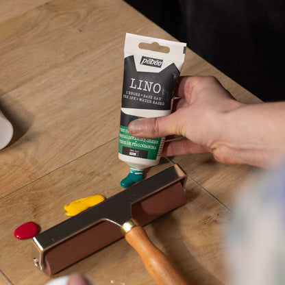 Person holding a tube of Pébéo Lino printing ink on a wooden surface with a printing roller and ink stones.
