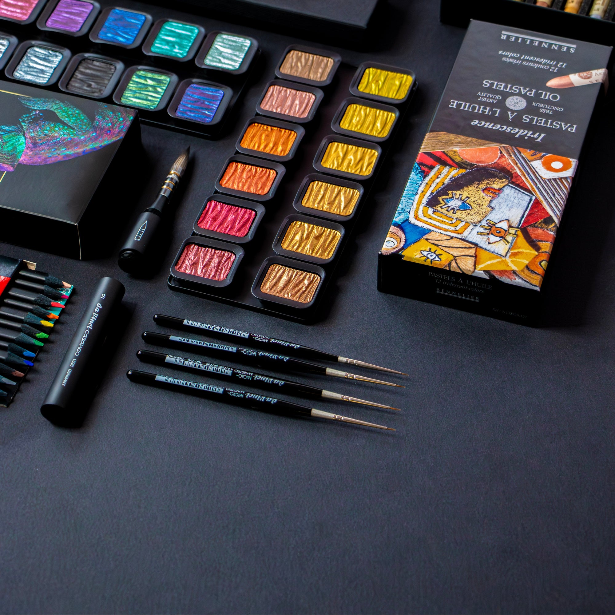 Collection of art supplies including watercolor paints, brushes, and books on a dark surface.